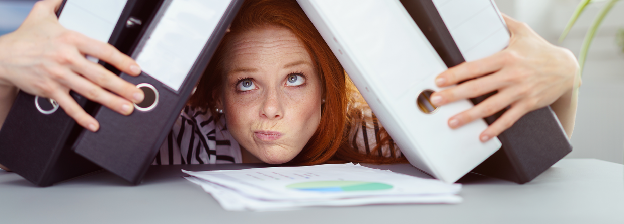 Burn-out; jonge vrouw met haar kin op een stapeltje papieren op haar bureau en houd ordners tegen haar hoofd aan